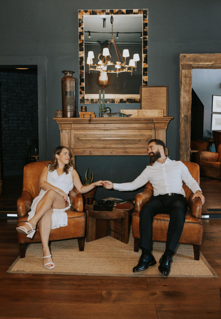 Couple seated in matching brown leather armchairs, reaching toward each other beneath a wooden fireplace mantel during their indoor engagement photos.