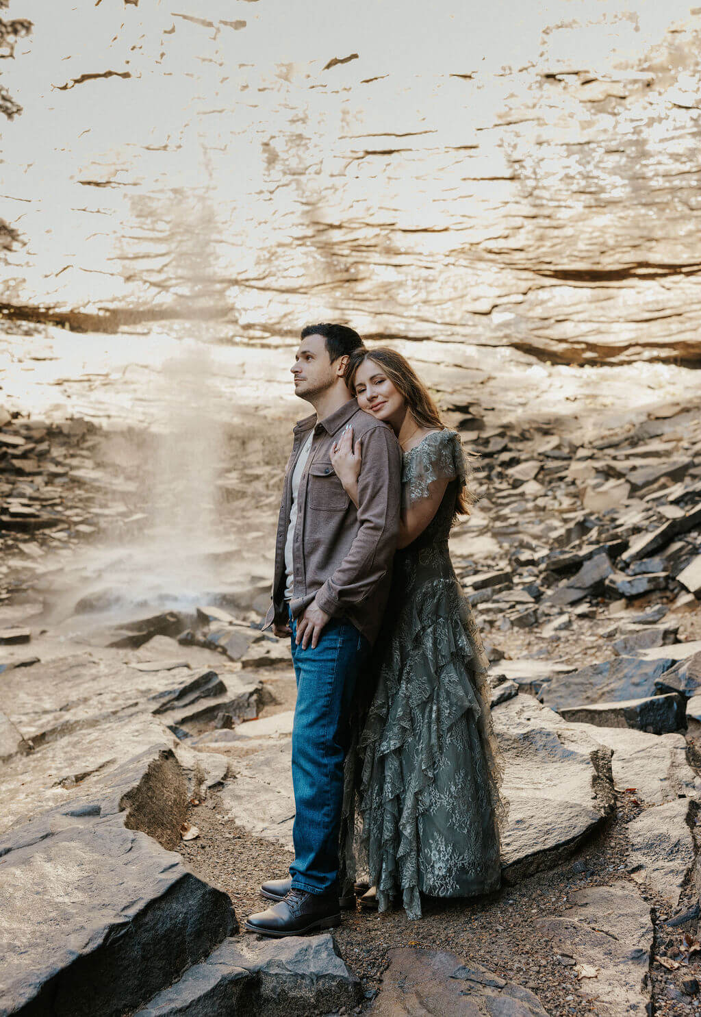 Couple posing together on rocky terrain in front of a waterfall during fall engagement photos.