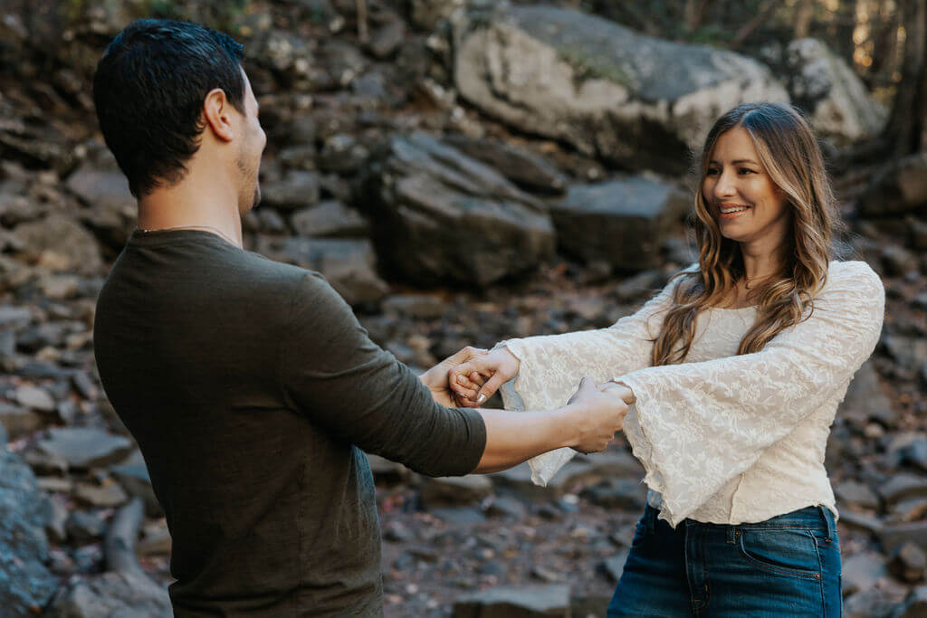 Couple laughing and holding hands on a rocky trail during playful fall engagement photos.
