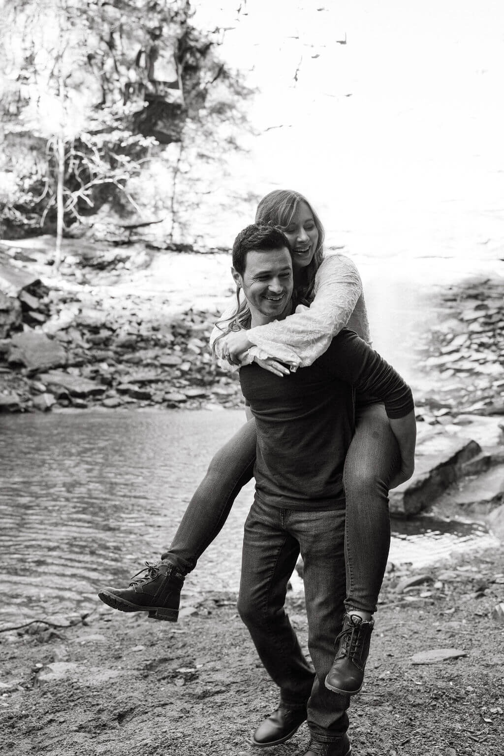 Black and white photo of a couple laughing during playful fall engagement photos by the water.