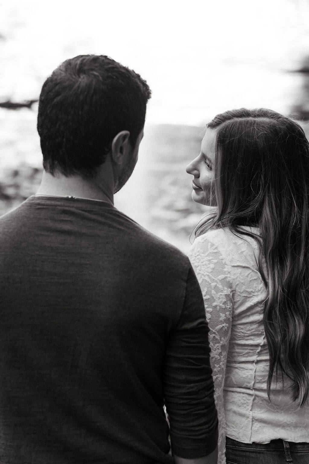 Black and white fall engagement photo of a couple smiling at each other beside the water.