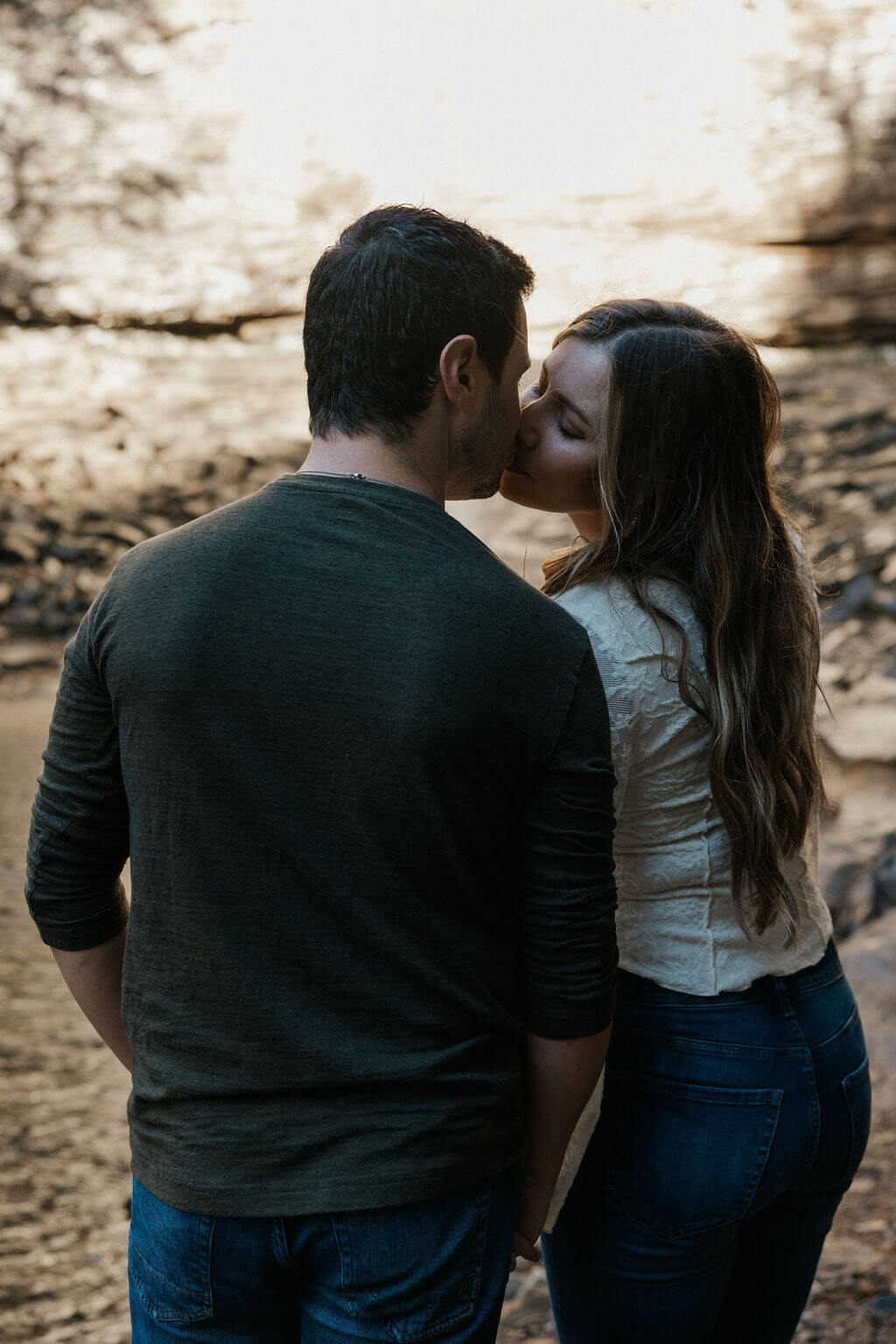 Couple sharing a quiet kiss beside the water during fall engagement photos with soft autumn light.