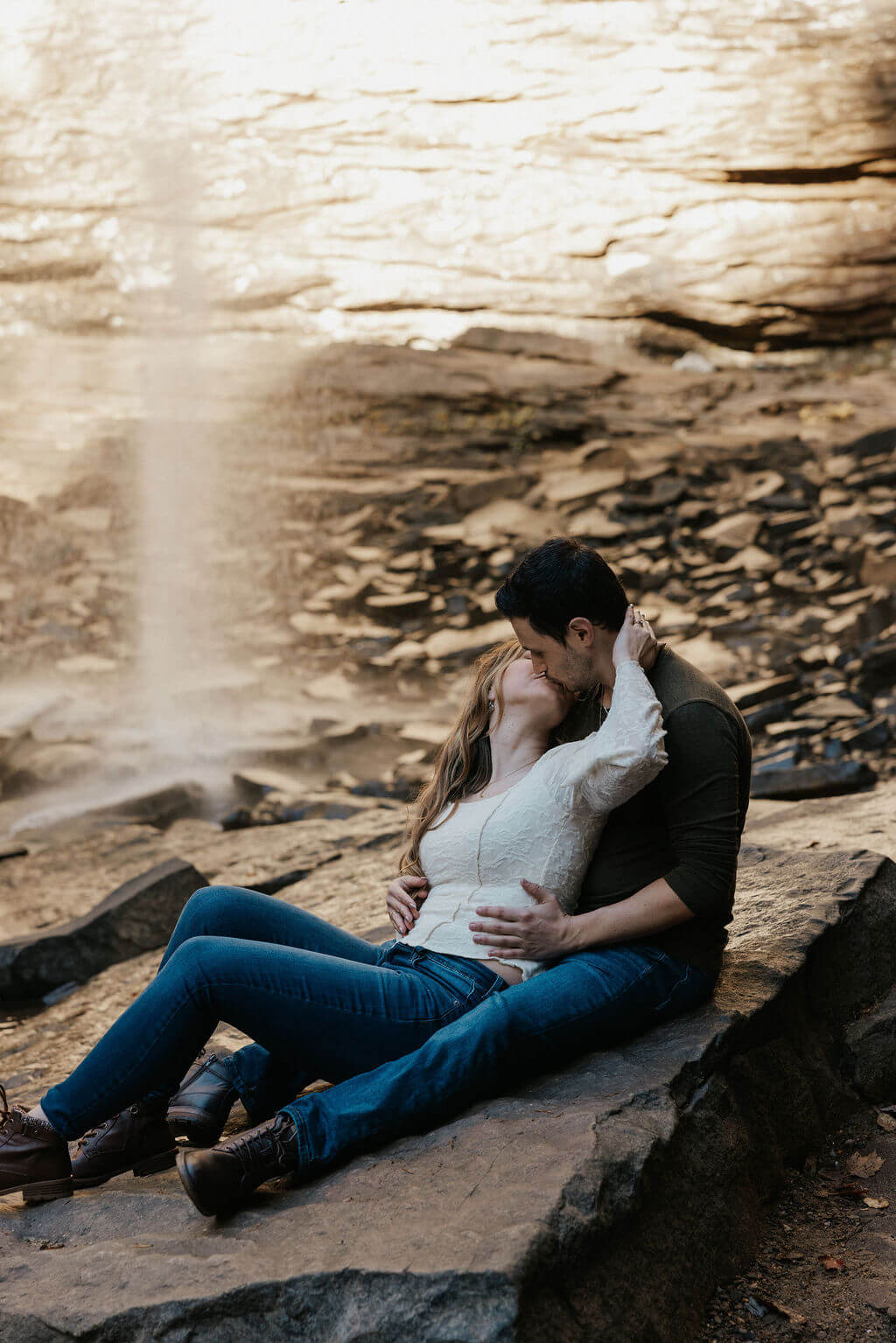 Couple sitting together on a rock and kissing in front of a waterfall during cozy fall engagement photos.