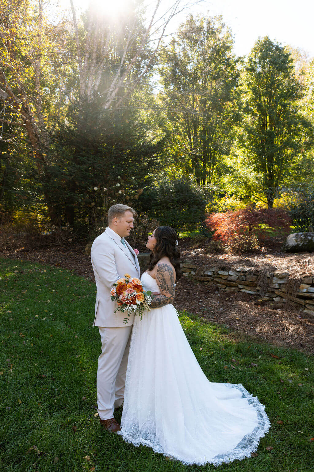 Couple stands together in a landscaped garden as sunlight streams through the trees at their fall wedding venue.