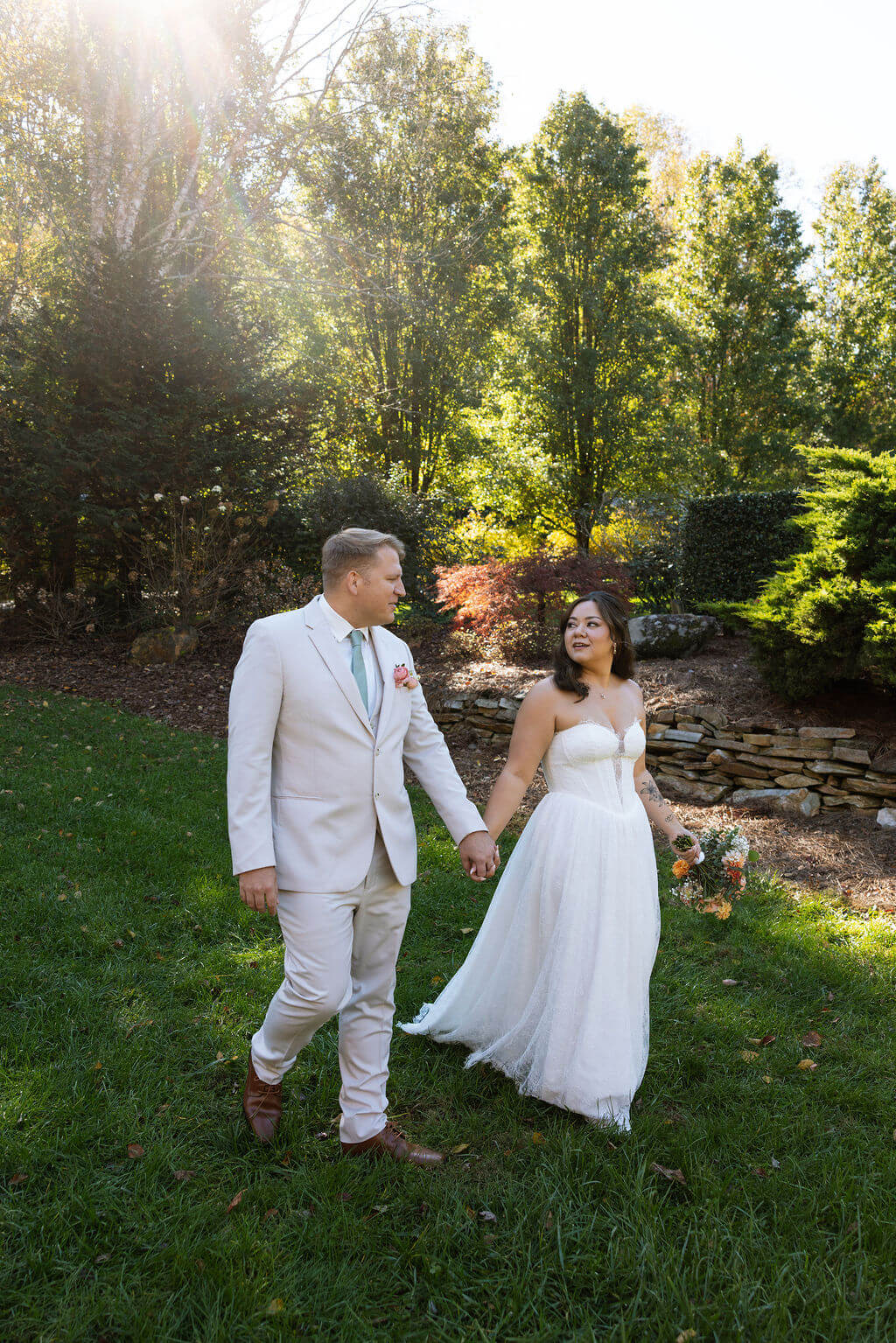 Newlyweds walk through a garden surrounded by green hedges, stone landscaping, and warm afternoon light.