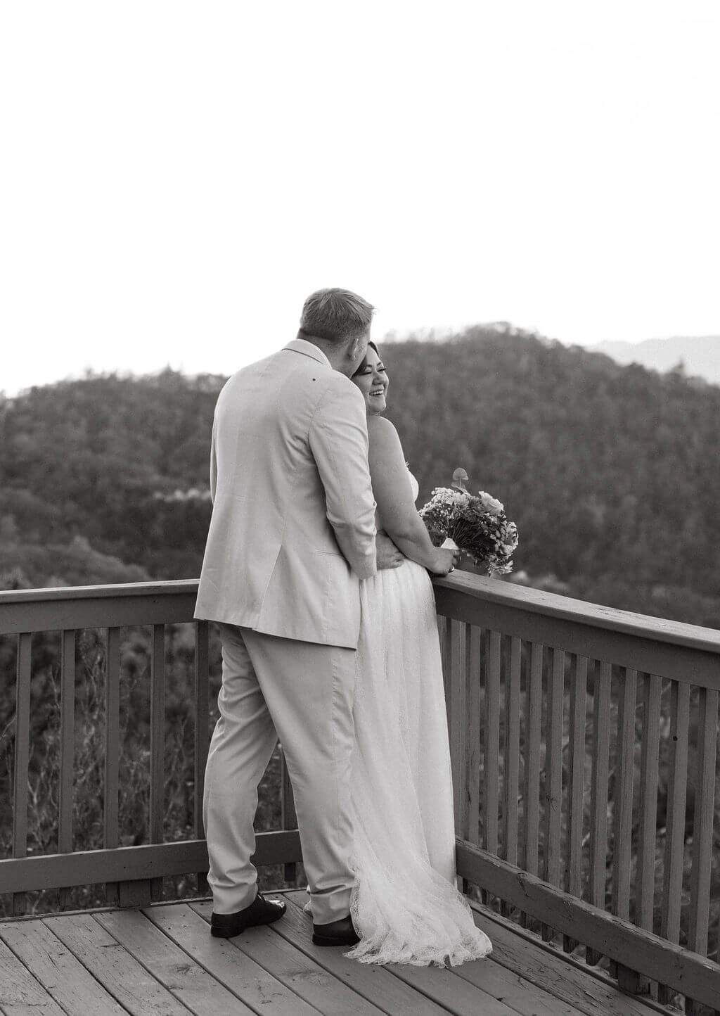 Black-and-white photograph of newlyweds kissing on a deck with mountain views behind them.