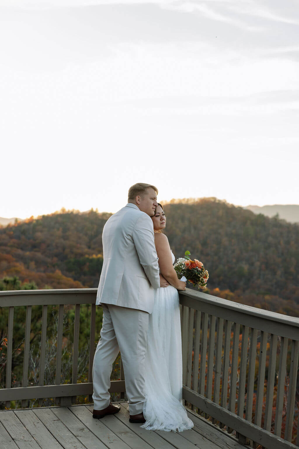 Newlyweds embrace on a cabin deck with rolling mountains and warm fall foliage in the background.