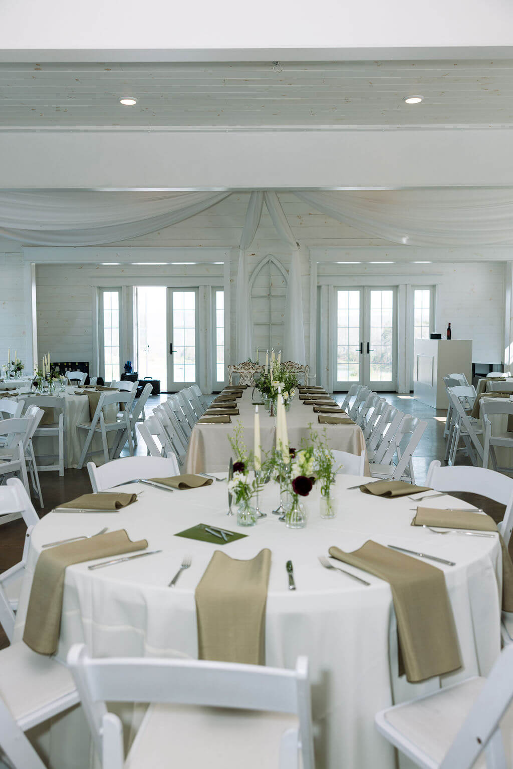 Bright wedding reception room decorated with round tables, white chairs, and neutral linens.