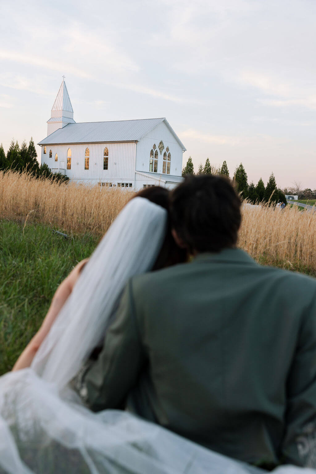 Newlyweds looking toward a white chapel while standing together in a grassy field.