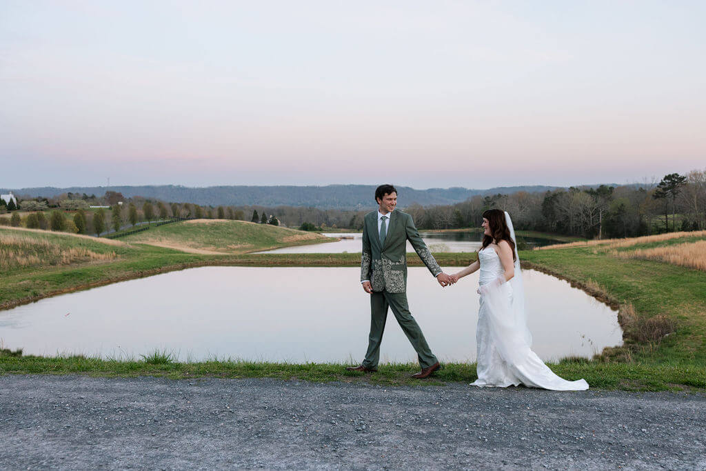 Bride and groom holding hands while walking beside a pond at a countryside wedding venue.