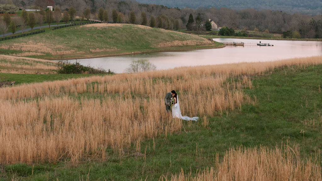Newlyweds walking through tall grass beside a pond at a countryside wedding venue.