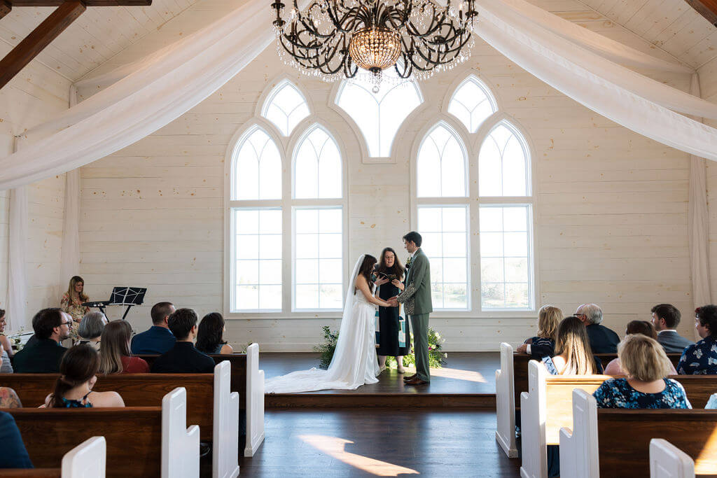 Couple exchanging vows inside a white chapel with arched windows and wooden pews.