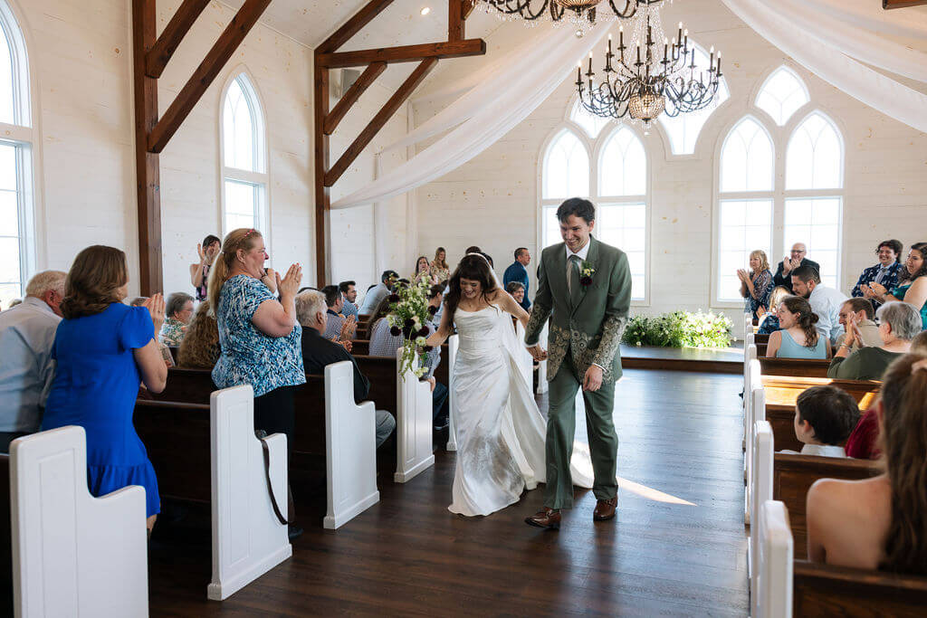 Newlyweds walking down the aisle after their ceremony inside a white chapel.