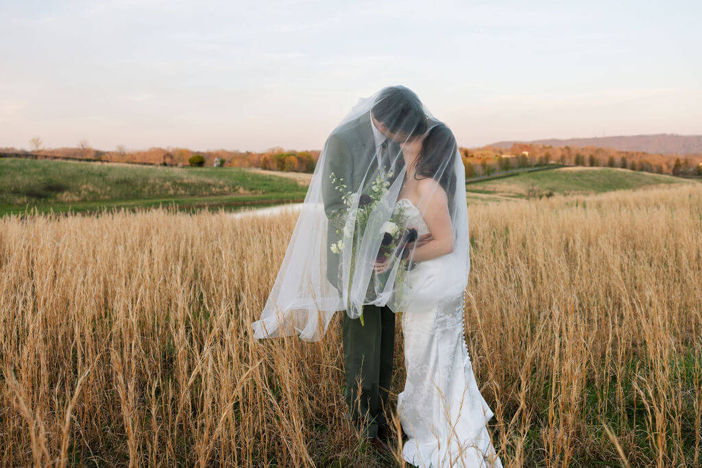 Newlyweds embracing in a grassy field at sunset beneath the bride’s veil.