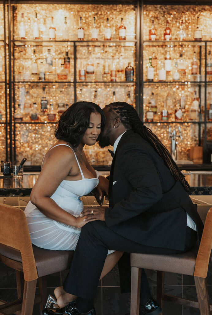 Engaged couple sitting close together in front of a glowing wall of liquor bottles during their romantic engagement photoshoot at a Knoxville bar.