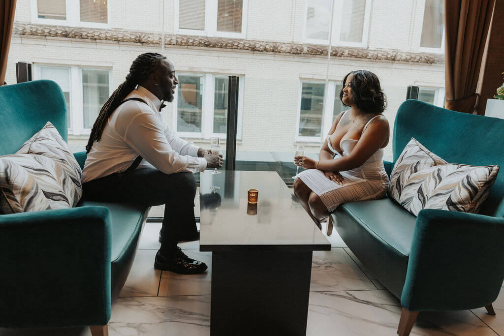 Couple in formal black-and-white outfits sitting across from each other on teal velvet sofas beside a large window during their engagement photoshoot.