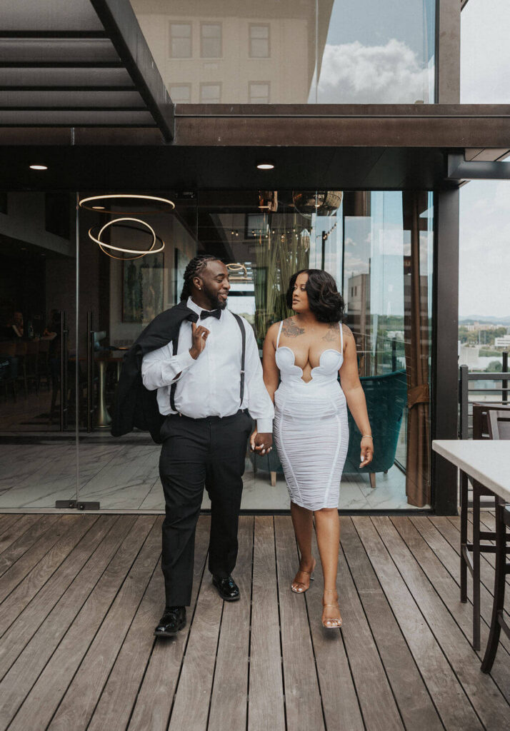 Couple in formal black-and-white outfits walking hand in hand across a modern rooftop patio during their engagement photoshoot at a Knoxville bar.