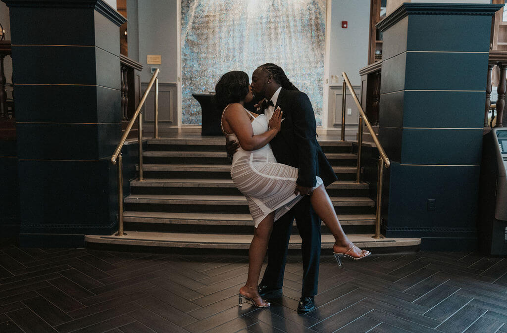 Engaged couple kissing in front of a grand staircase, with the woman lifting one leg during their playful indoor engagement photoshoot.