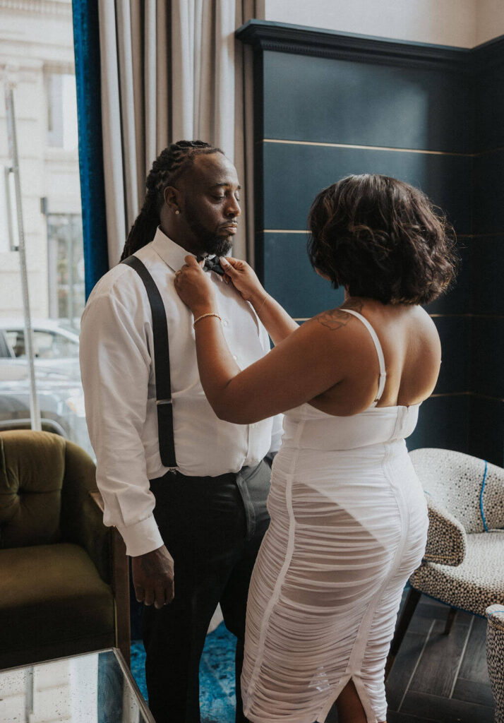 Woman adjusts her fiancé’s bow tie as he wears black suspenders during an elegant engagement photoshoot inside a Knoxville bar.