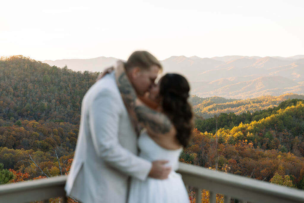 Softly focused newlyweds kiss on a deck overlooking expansive Smoky Mountain fall foliage.
