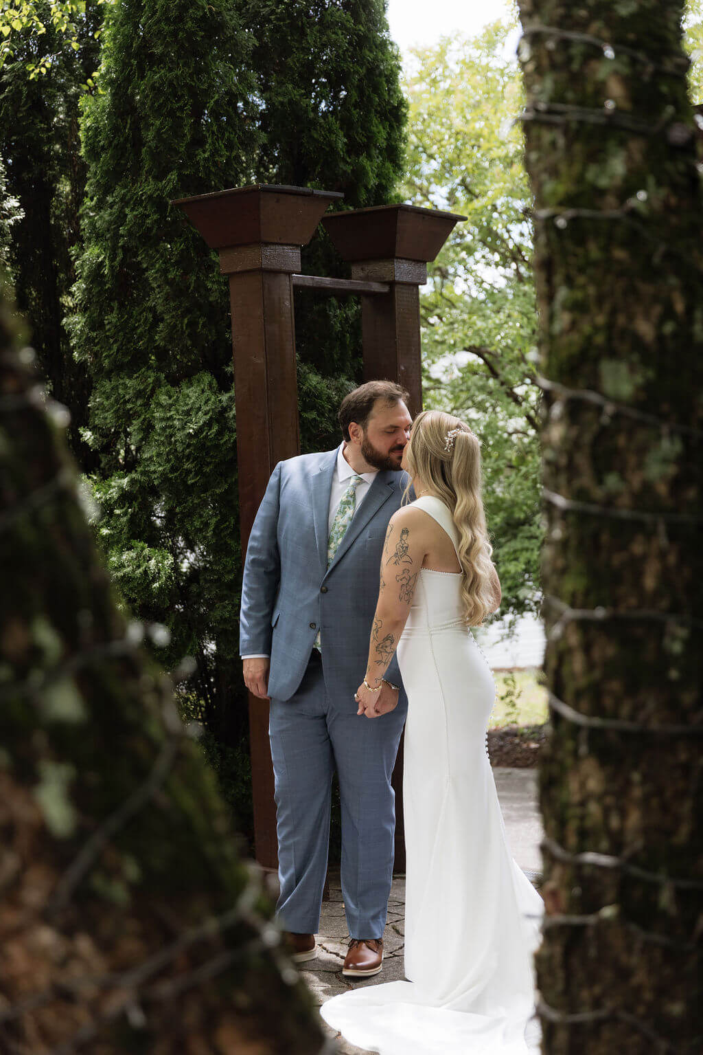 Bride and groom kissing beneath a wooden arbor at a landscaped wedding venue.