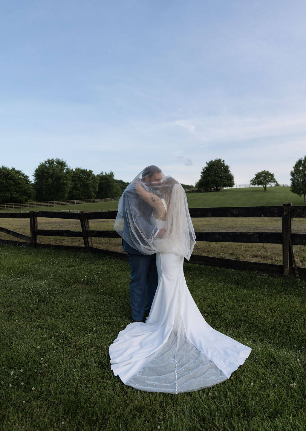 Bride and groom embracing beneath the bride’s veil beside a wooden pasture fence.