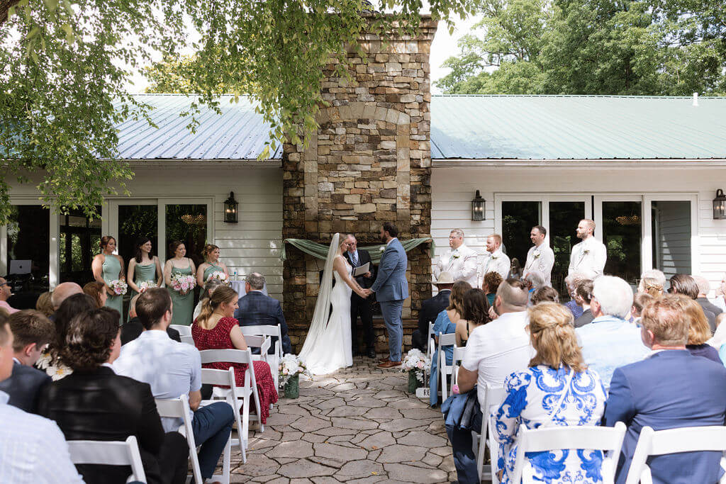Couple exchanging vows during an outdoor wedding ceremony beside a stone building.