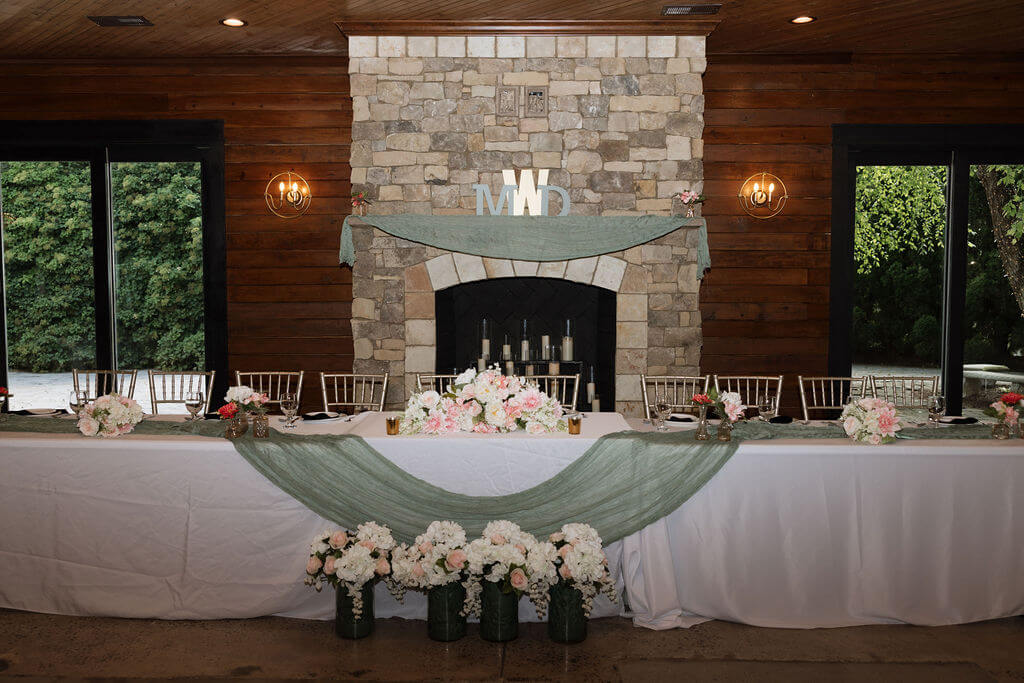 Wedding sweetheart table decorated with flowers and draped fabric in front of a stone fireplace.