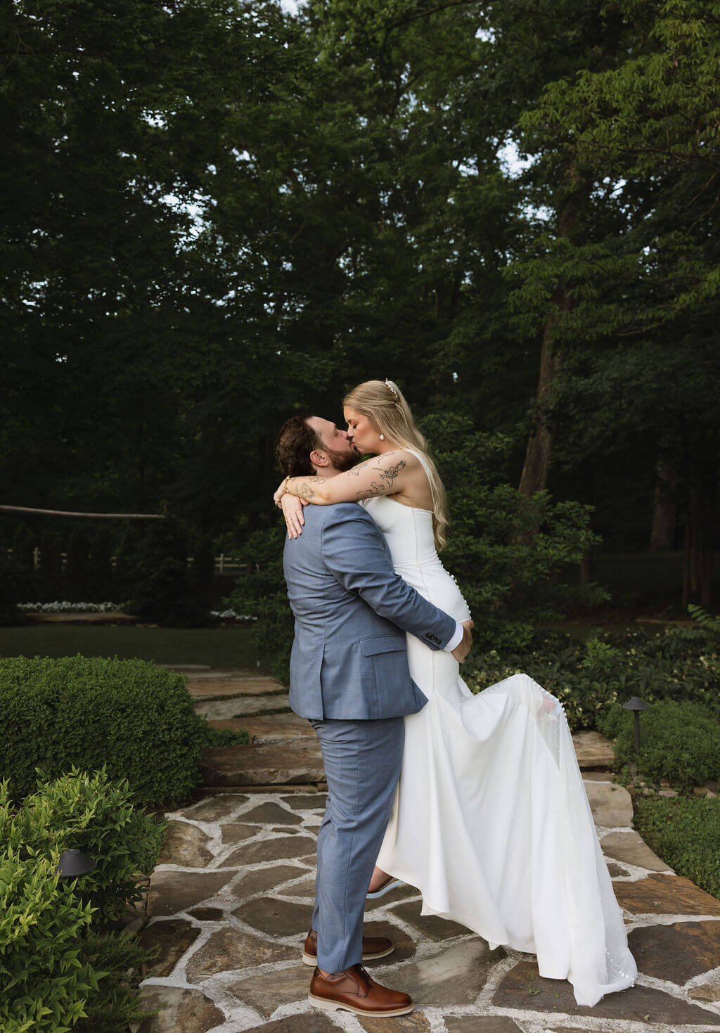 Groom lifting and kissing the bride on a stone pathway surrounded by lush trees and gardens.