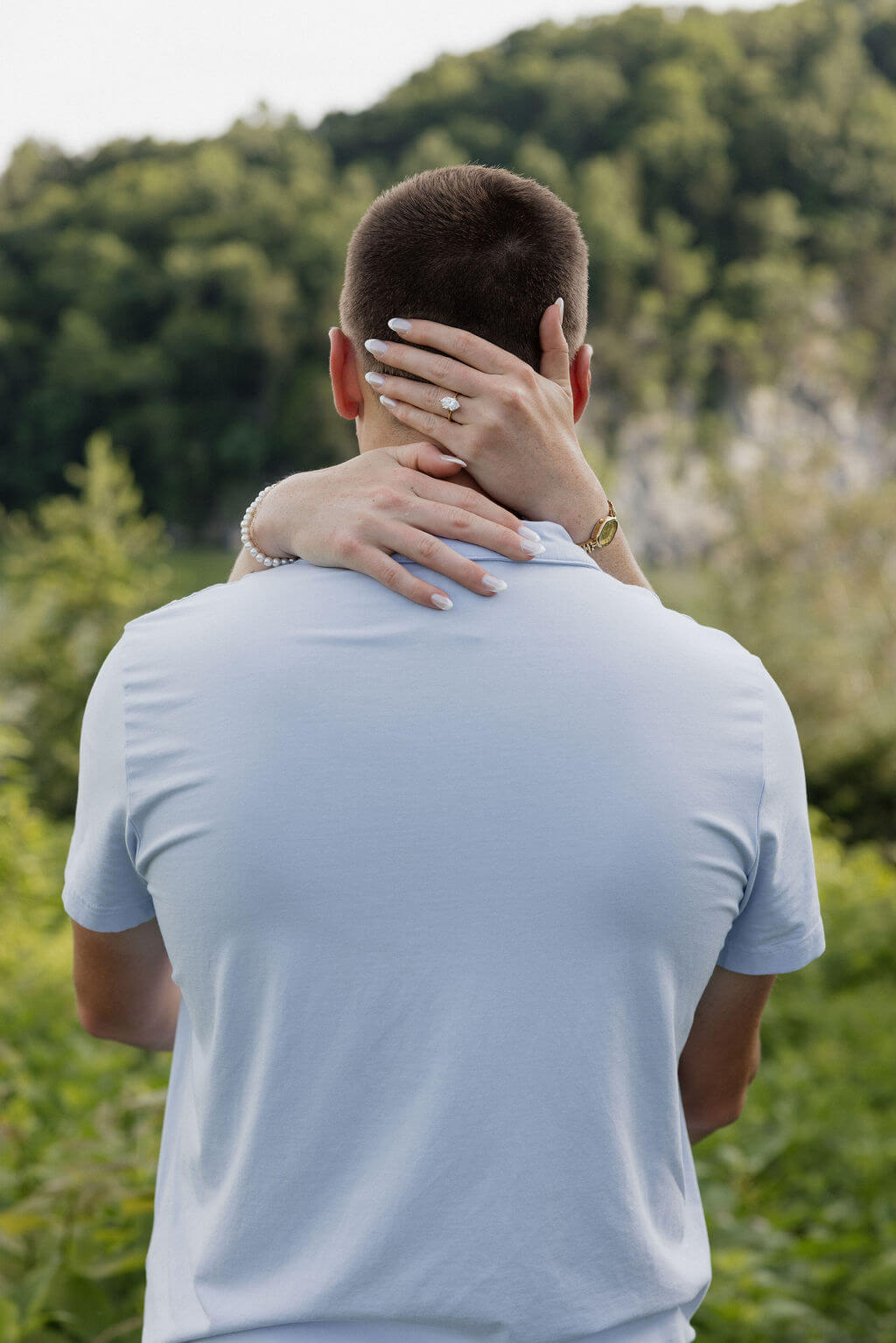 Woman wrapping her arms around her fiancé beside a lake and wooded hillside.