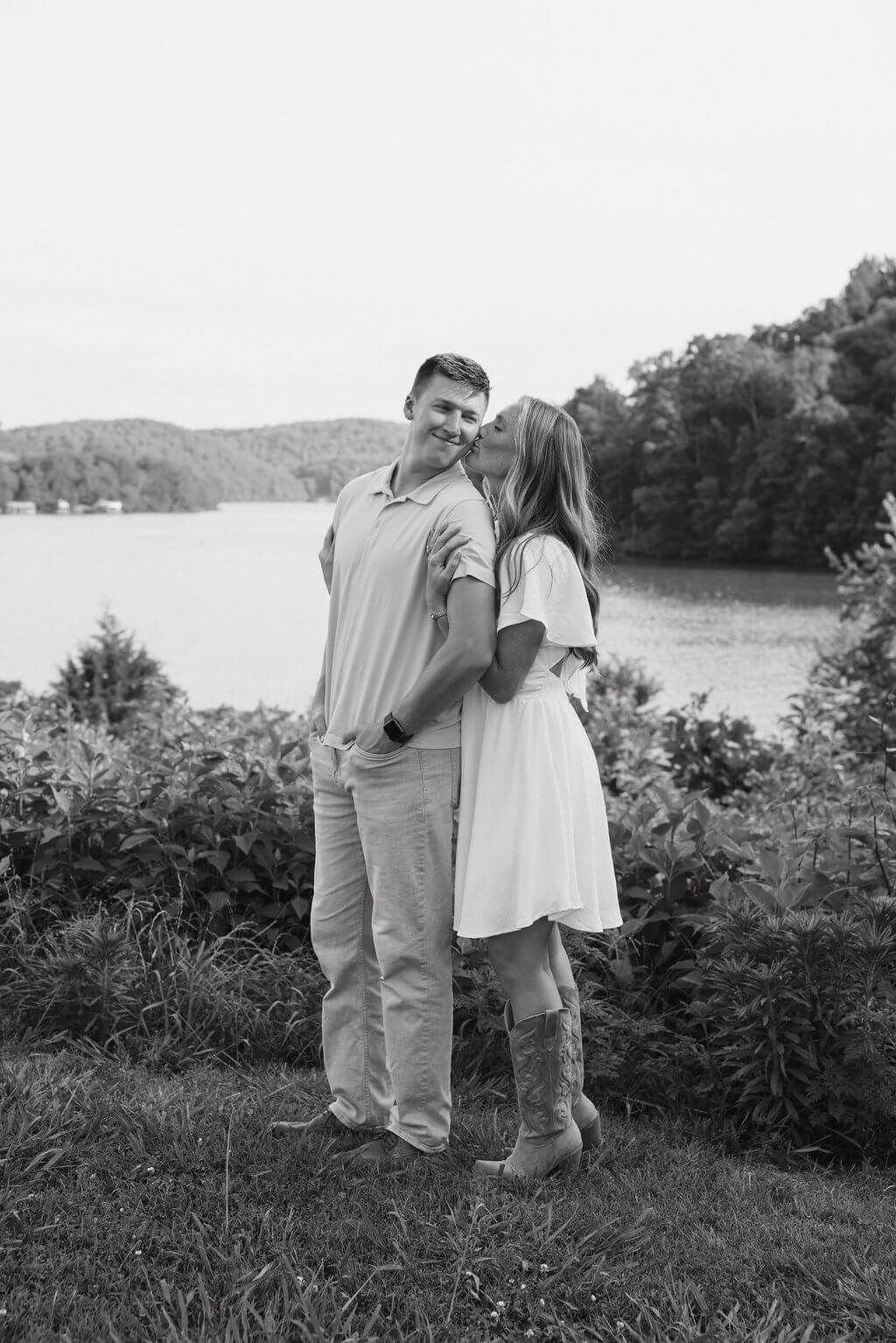Black-and-white portrait of an engaged couple standing together beside the lake.