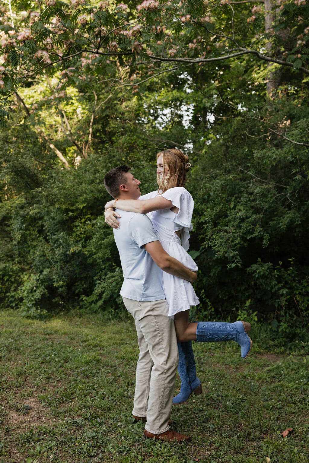 Man lifting his fiancée as they laugh together beneath leafy trees.