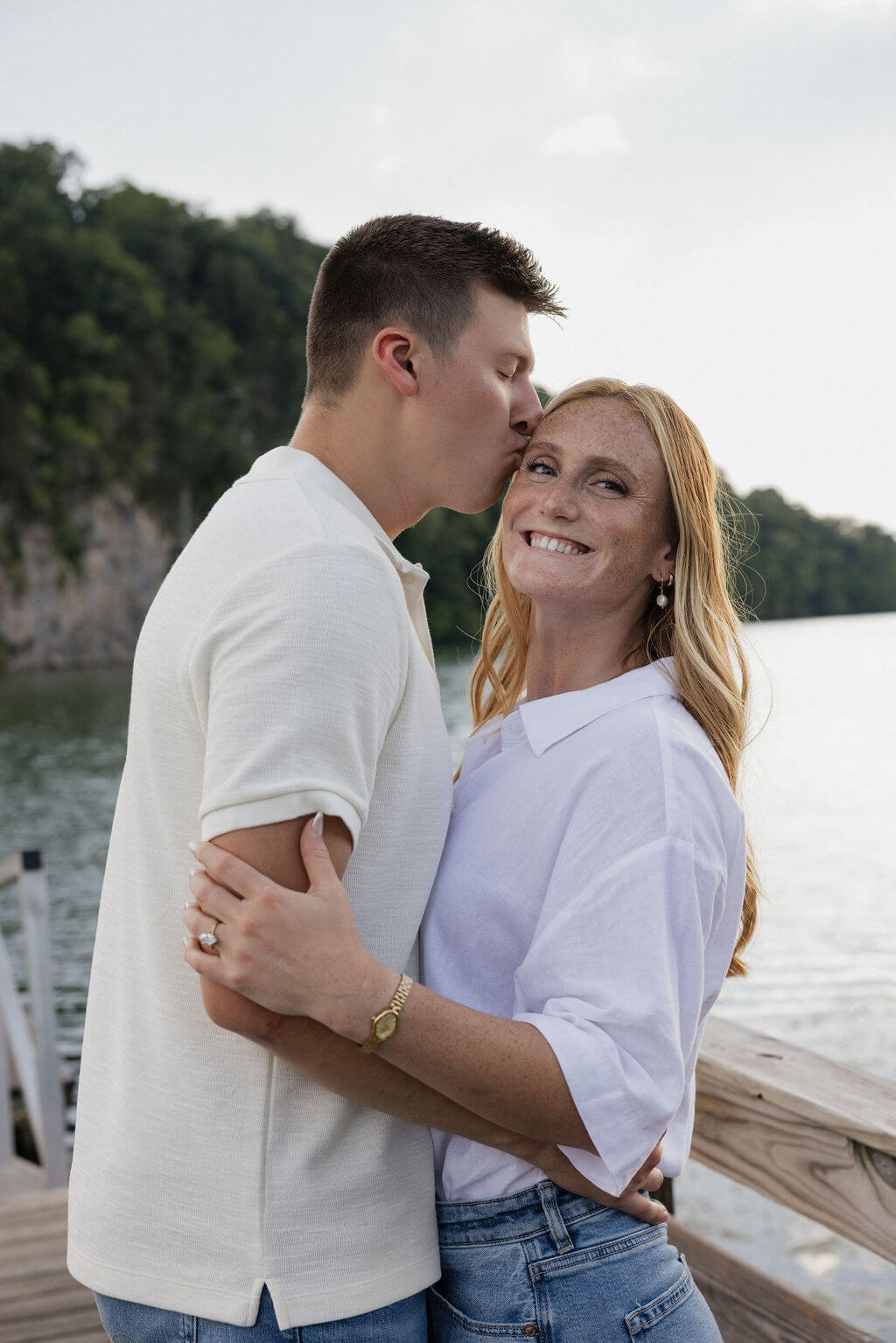 Man kissing his smiling fiancée on the cheek while they stand on a wooden dock.