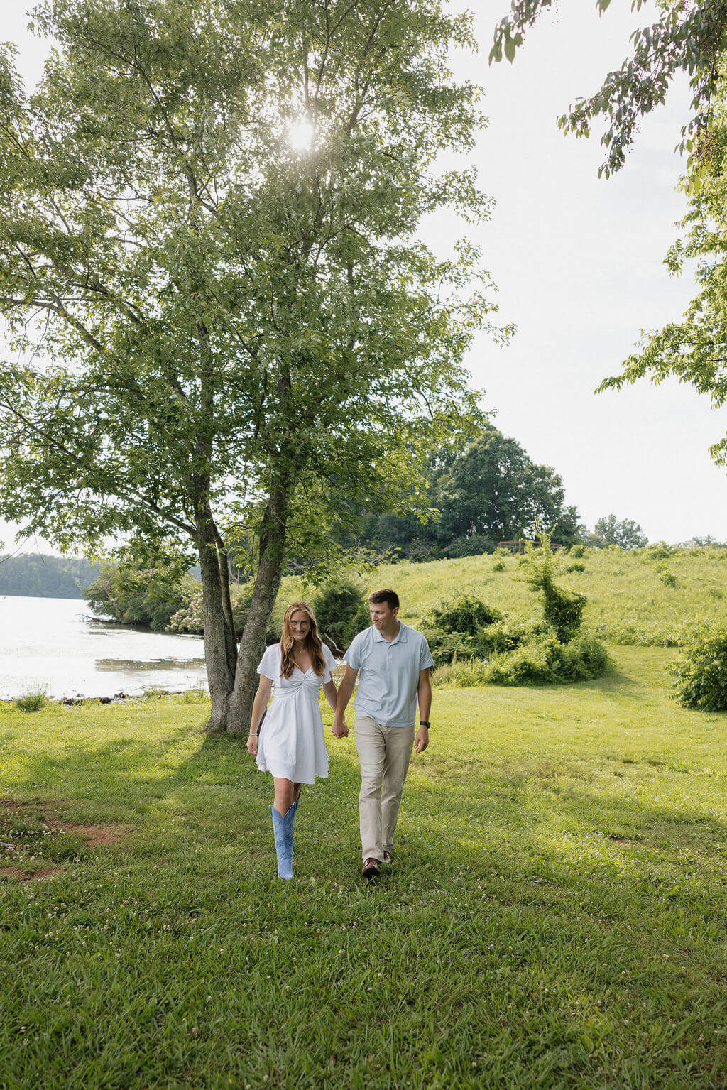 Engaged couple walking hand in hand beneath a large tree beside the water.