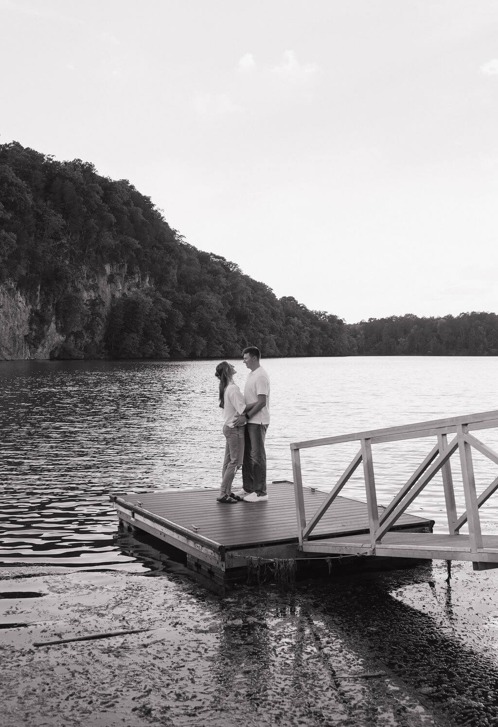 Black-and-white photo of an engaged couple embracing on a dock overlooking the water.