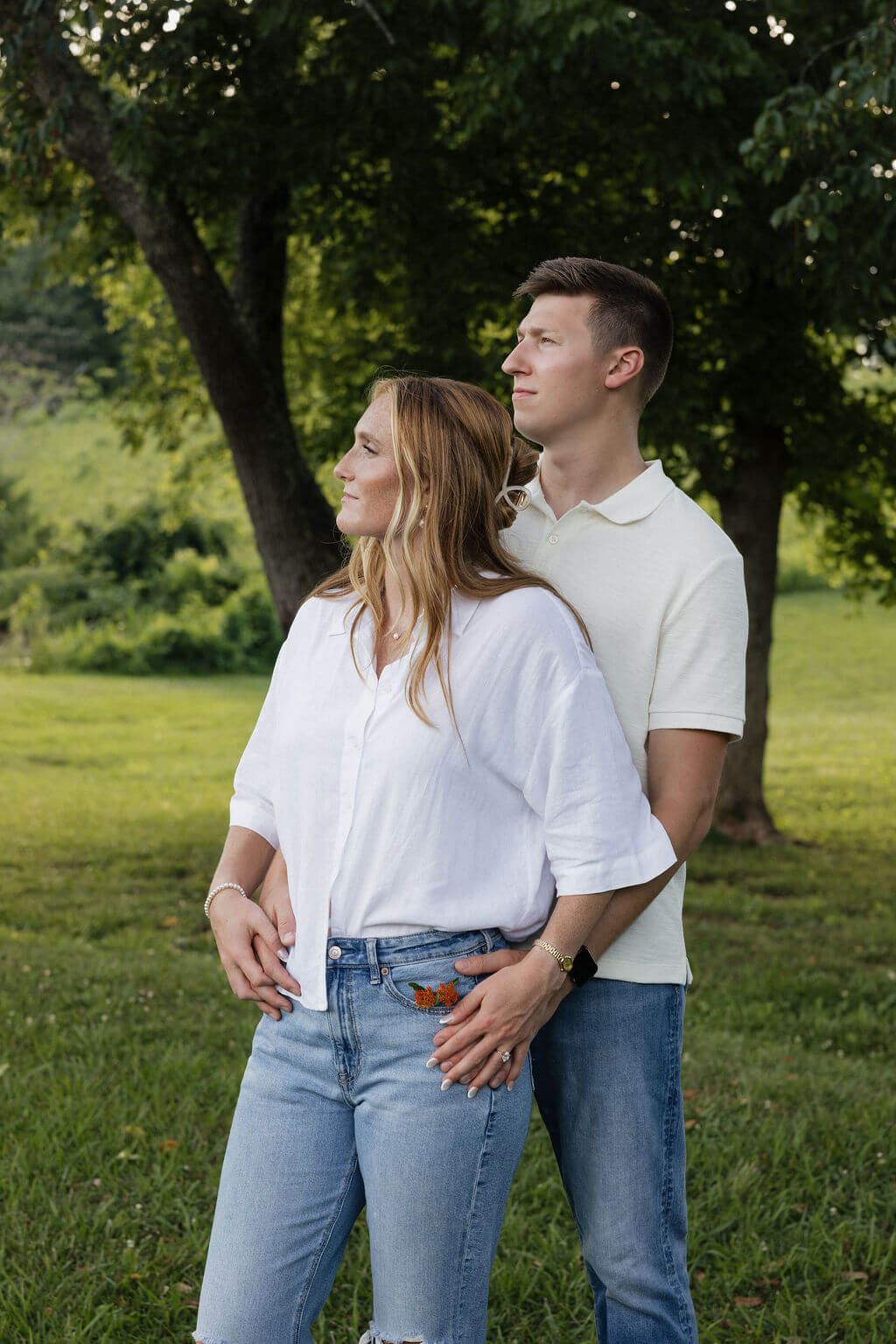 Engaged couple standing together beneath a shady tree near the water.