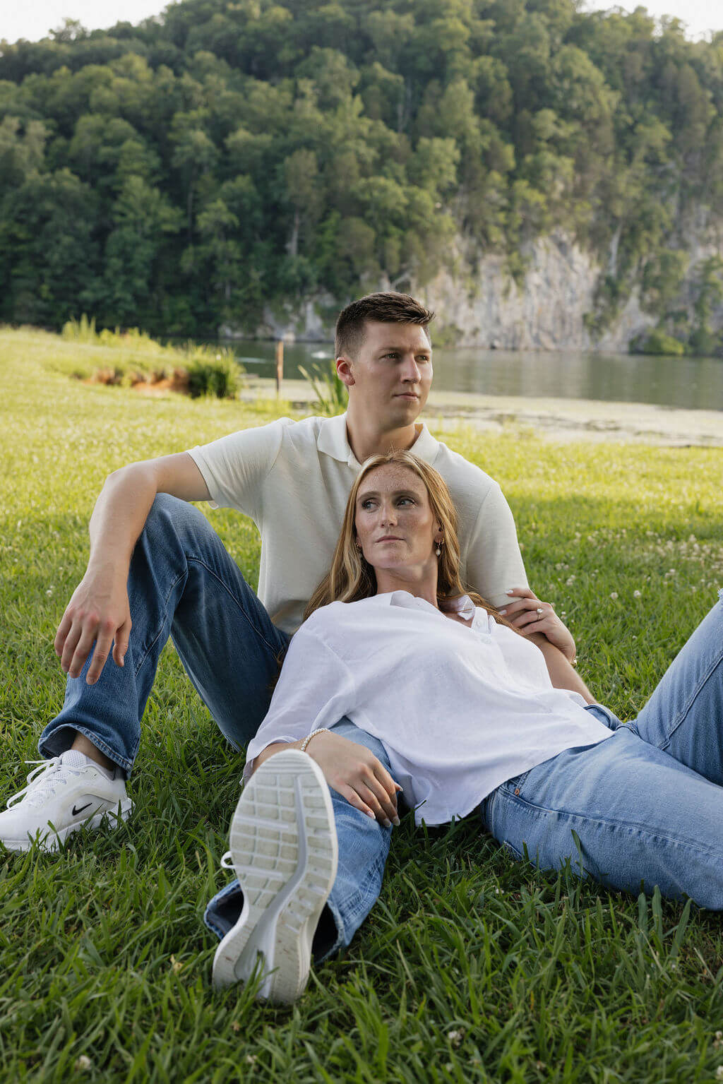 Couple relaxing in the grass with a lake and rocky wooded cliff behind them.