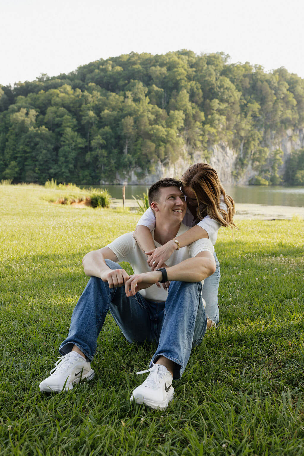 Engaged couple sitting close together and laughing beside the lake.