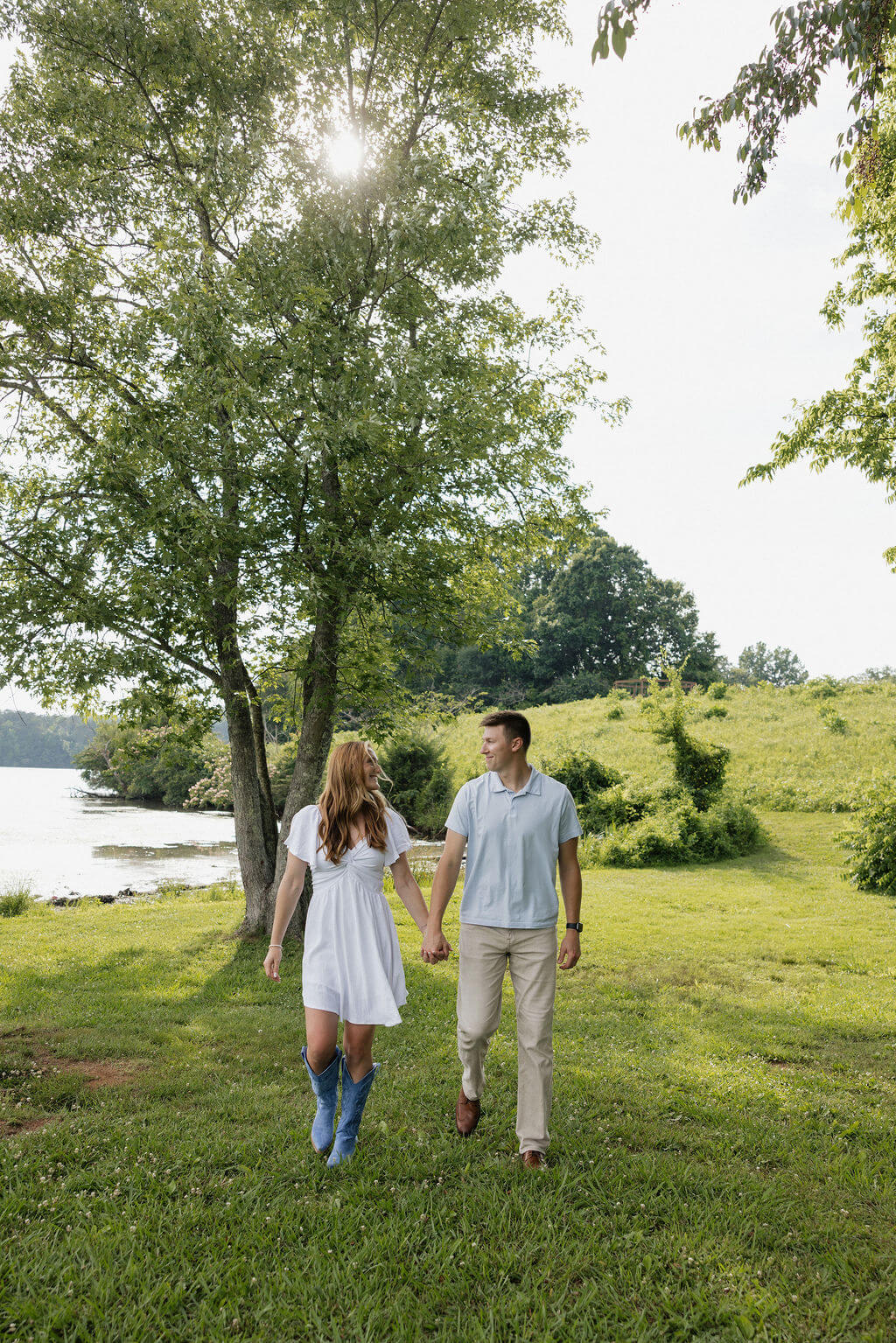 Couple walking hand in hand through a grassy lakeside setting beneath tall trees.