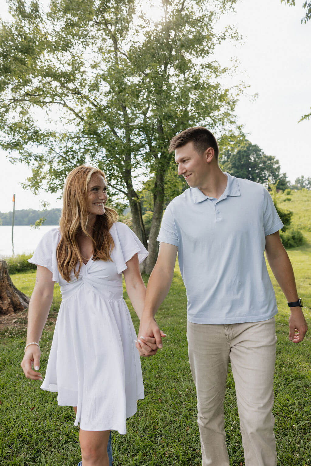 Engaged couple holding hands and smiling while walking beside the lake.
