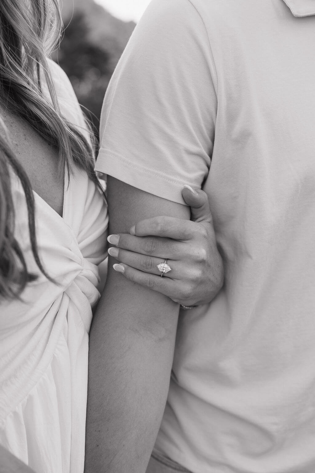 Black-and-white close-up of a woman’s engagement ring as she holds her partner’s arm.