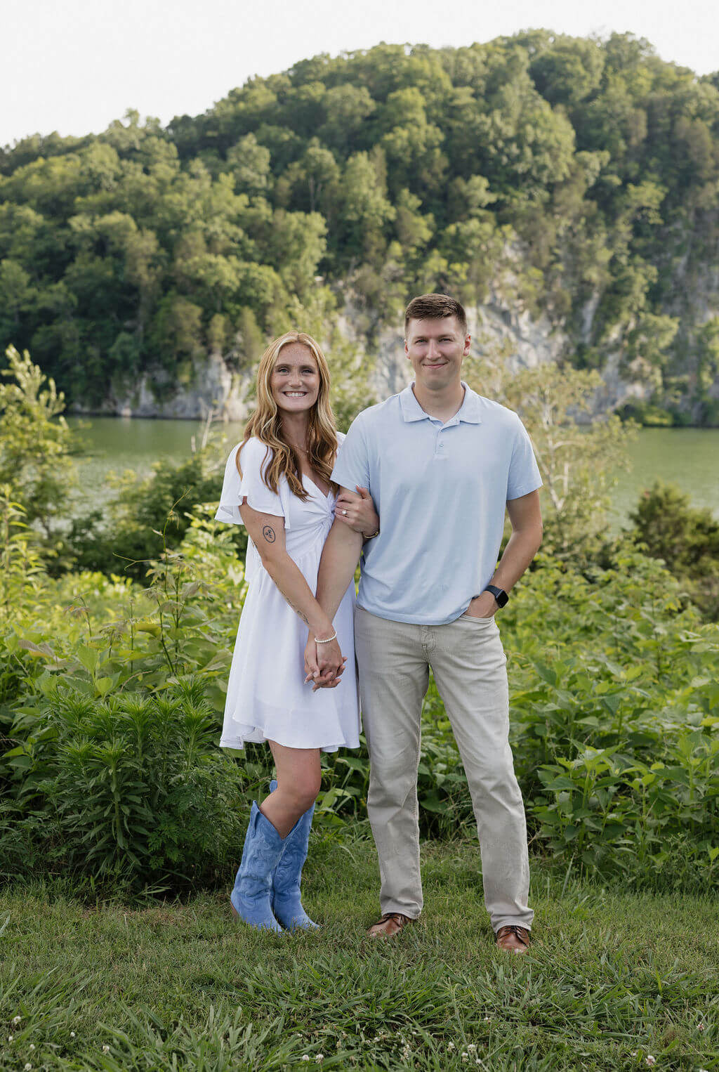 Couple posing together in front of a wooded cliff and calm lake.