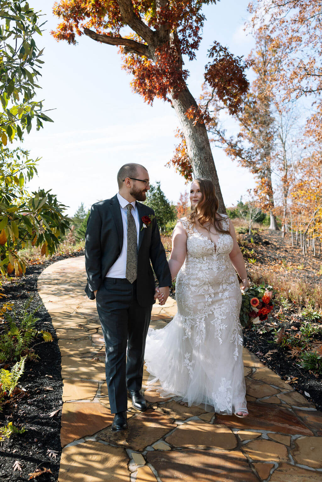 Newlyweds hold hands while walking along a stone garden path surrounded by trees and fall landscaping.