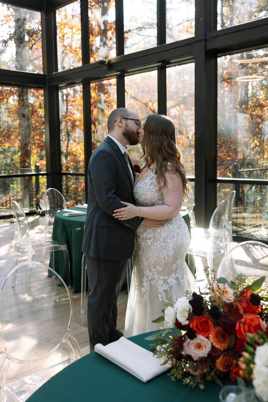 Couple embraces inside a glass-walled reception space decorated with green linens and autumn wedding flowers.