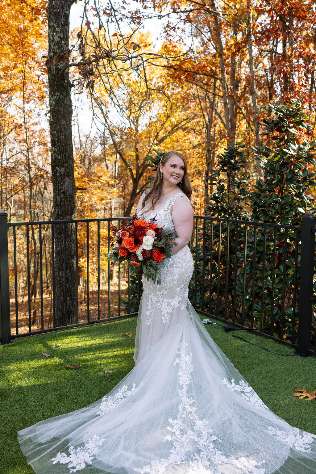 Bride holds a red and orange bouquet beneath a canopy of glowing fall leaves.