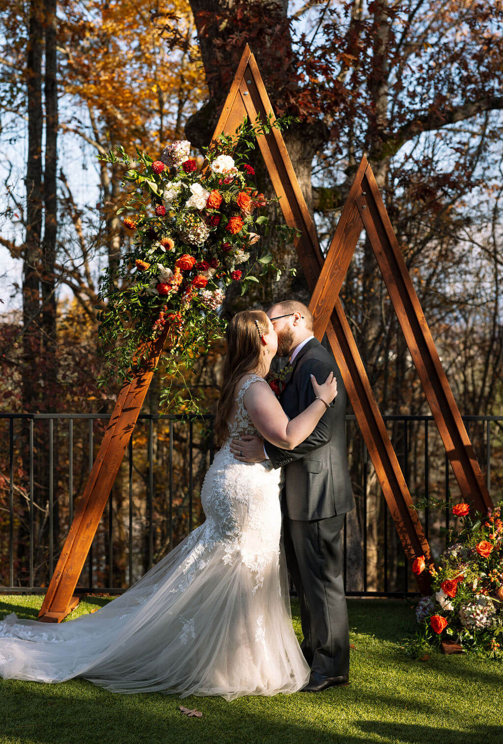 Newlyweds kiss beneath a wooden geometric ceremony arch decorated with red, orange, and white flowers.