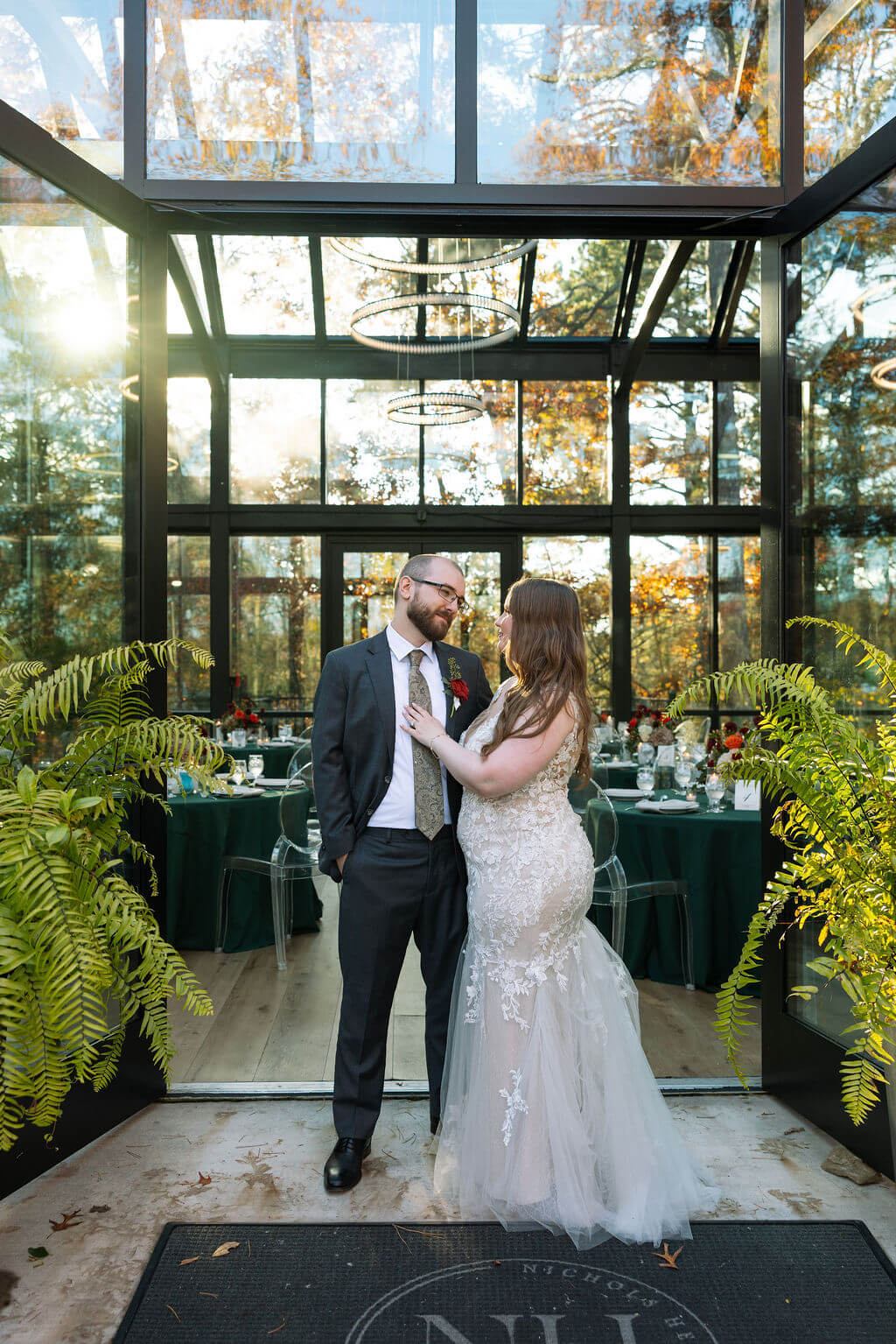 Couple embraces outside a glass-walled reception venue decorated with greenery and deep green linens.