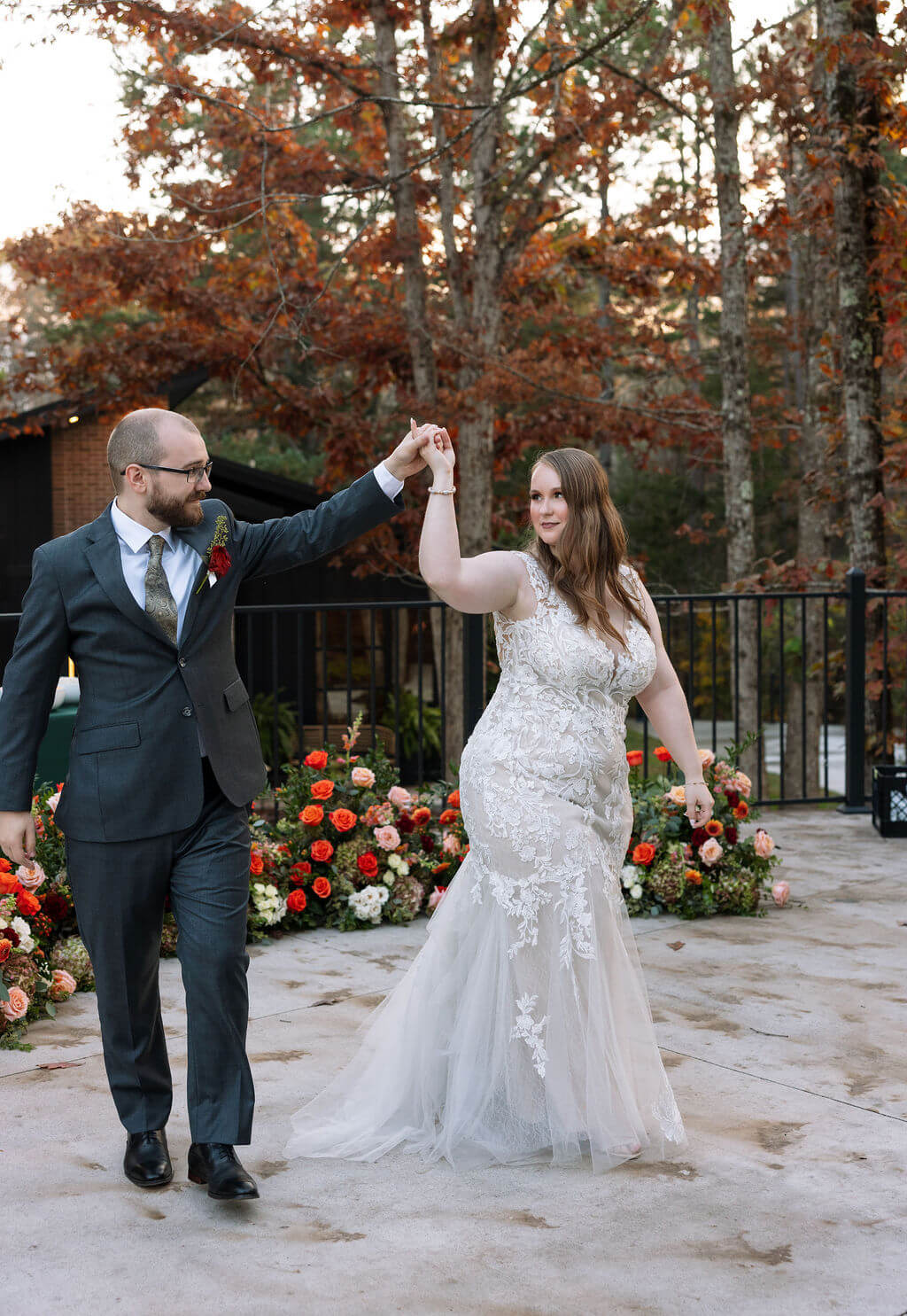Newlyweds dance on a stone patio surrounded by red flowers and autumn trees.