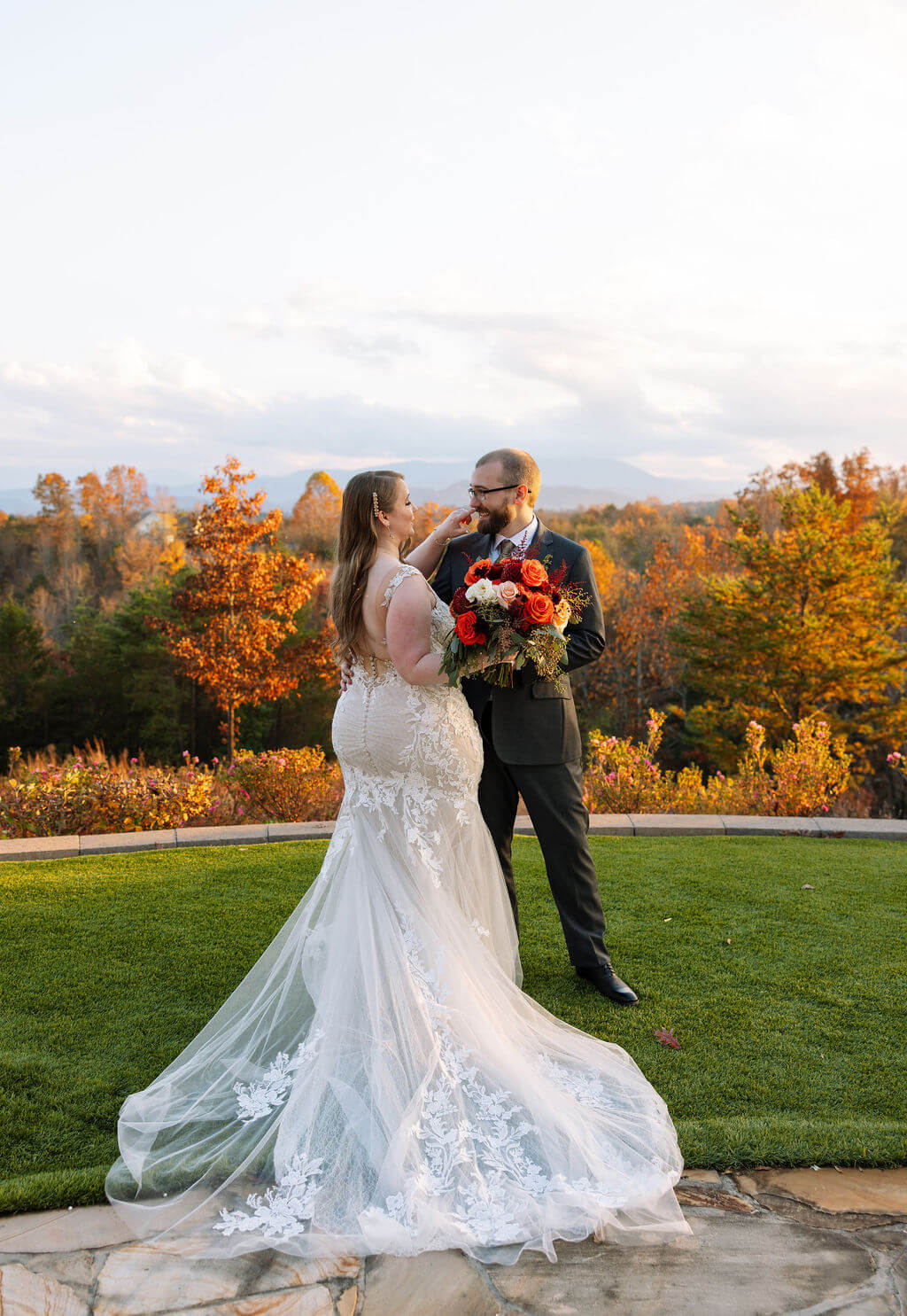 Newlyweds stand together with the bride’s lace train spread across the lawn and fall mountains behind them.