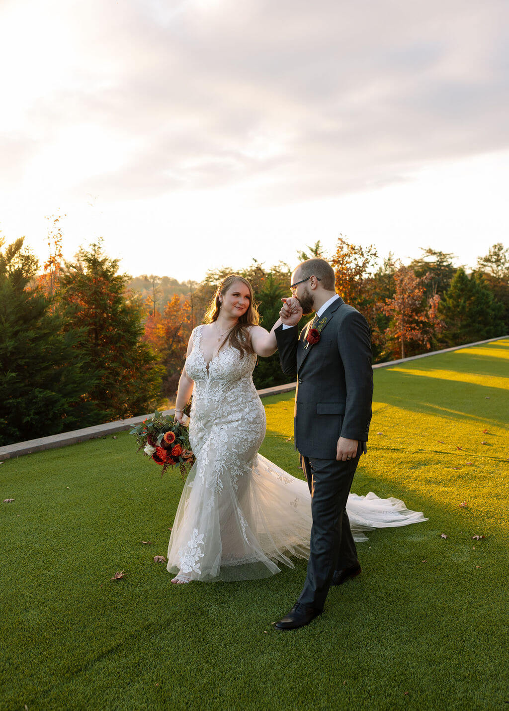 Newlyweds walk across a green lawn surrounded by colorful fall trees and distant mountain views.